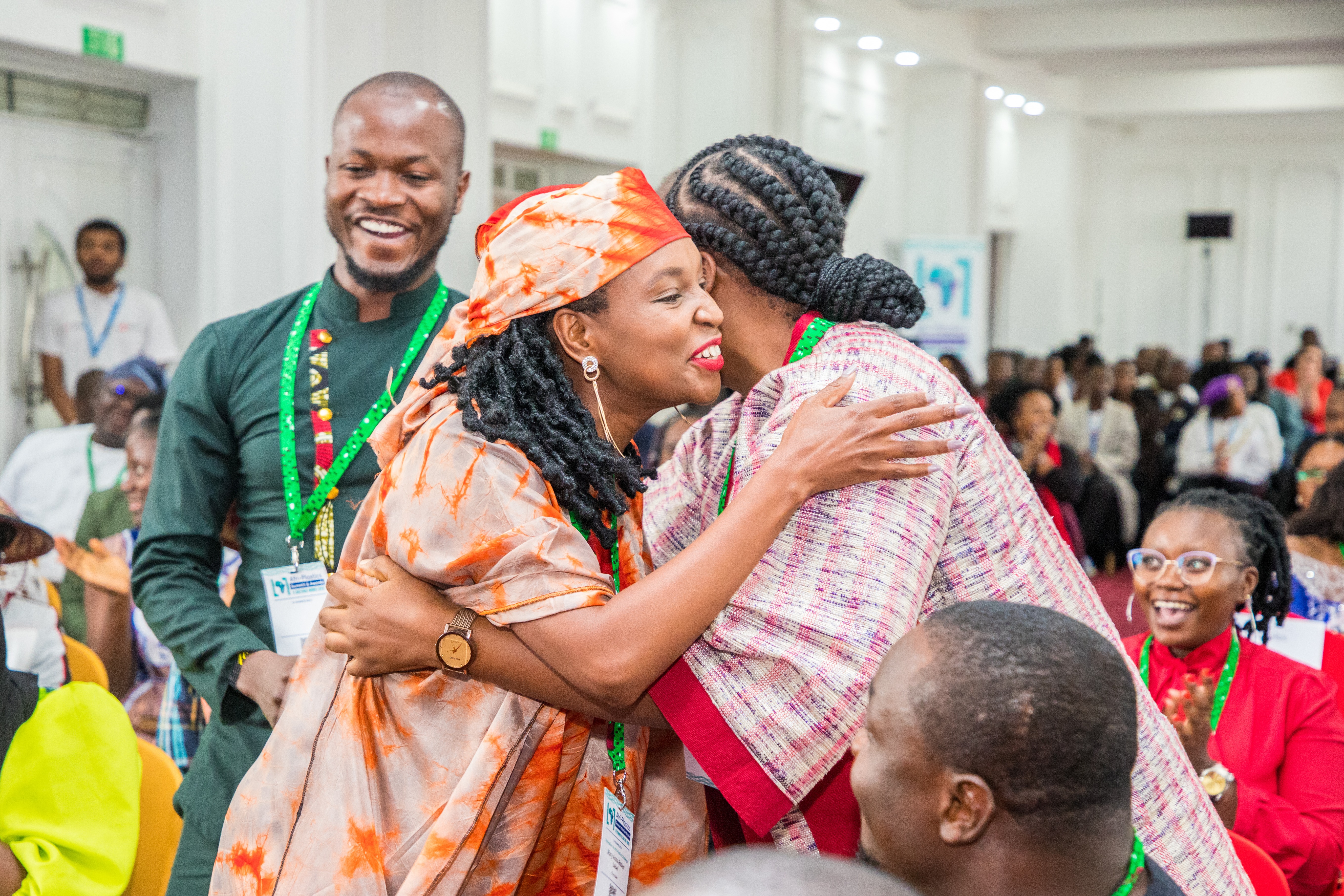 A joyful moment at a challenge prize celebration where two women are warmly embracing in a hug. One woman wears a vibrant orange and cream tie-dye garment with a matching headwrap and a wristwatch, while the other wears a patterned pink textured outfit and has her hair styled in thick braids. Surrounding attendees, including a man in a dark green tunic with a green lanyard, look on with bright smiles.