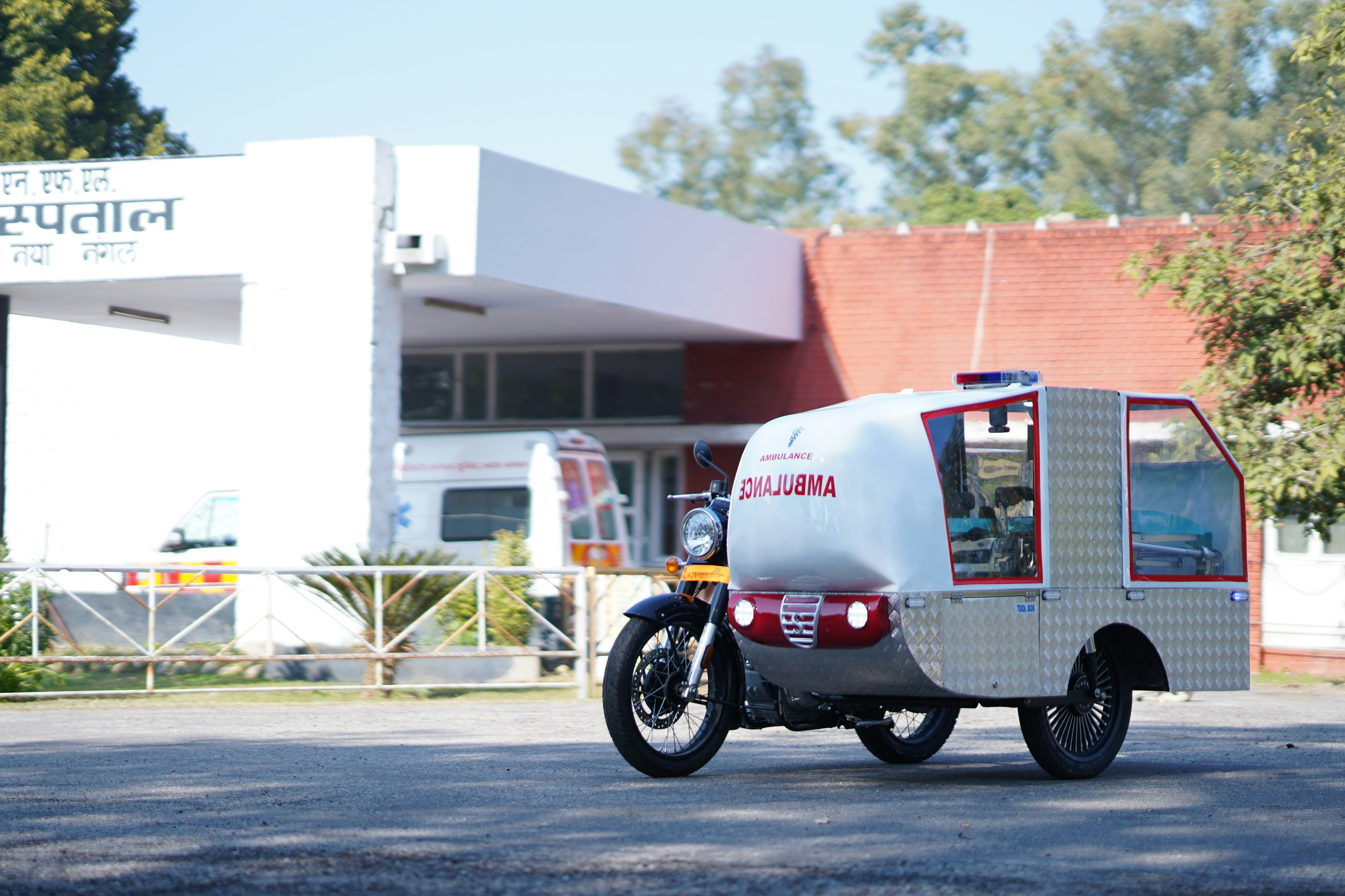 A specialised three-wheeled motorised ambulance vehicle parked outside on a driveway. The vehicle features a motorcycle front end attached to a silver, diamond-plate enclosed cabin with red trim, clear glass windows, and an emergency light bar on the roof.